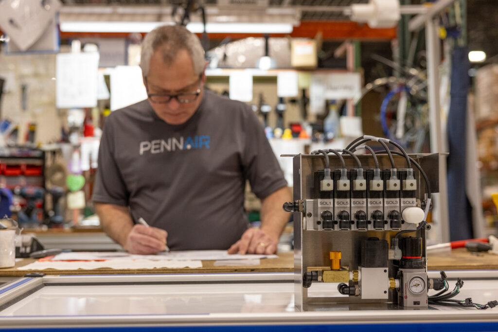 Man in a workshop signs a sheet at a workbench, with a hydraulic control panel in the foreground.