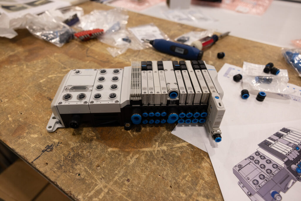 Close-up of a modular industrial automation controller with blue circular connectors on a workbench, surrounded by tools and bags of parts.
