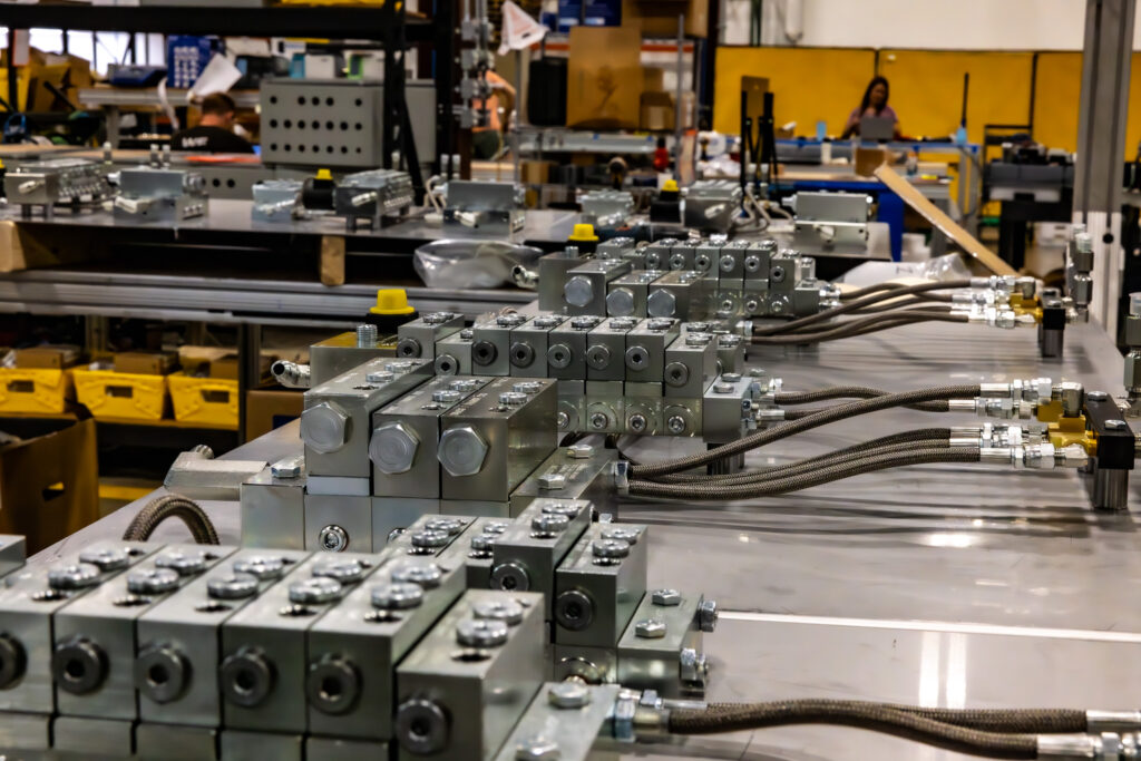 Close-up of gray hydraulic manifolds and fittings on a workbench in a factory setting.