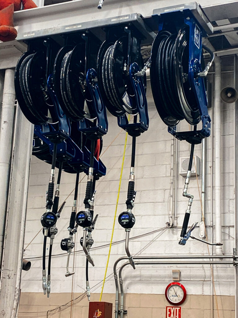 Row of blue cable reels with black hoses hanging from a metal rack in an industrial room with visible gauges and wiring