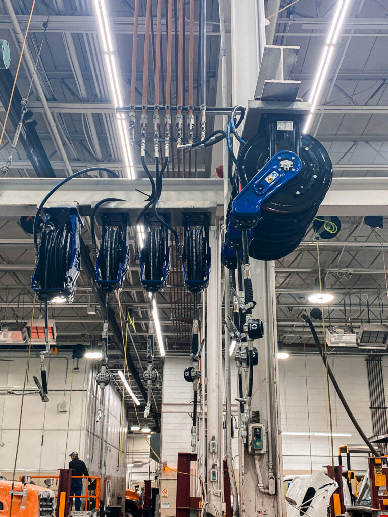 Industrial workshop scene with ceiling-mounted reels, hoses, and a large blue hoist/arm overhead.