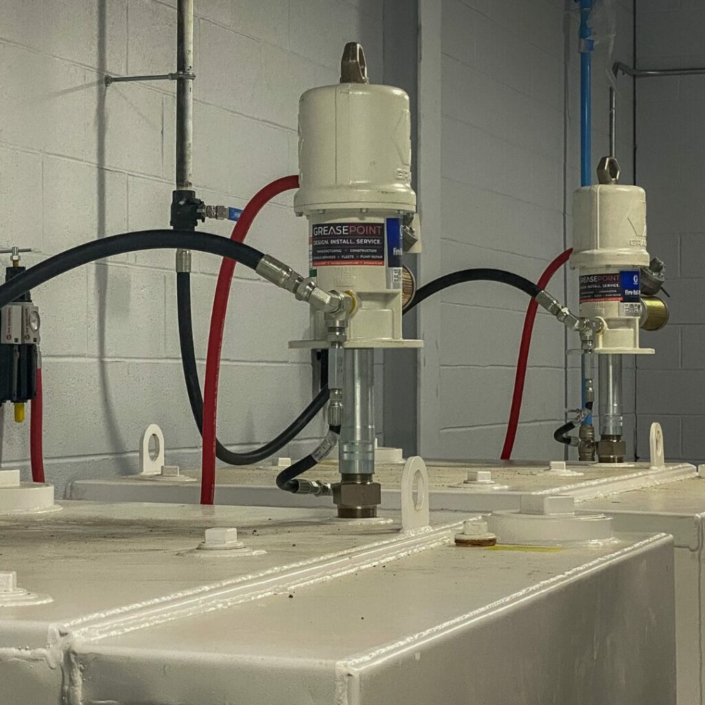 Two white grease-pump units with red and black hoses connected, mounted on white industrial tanks in a workshop.